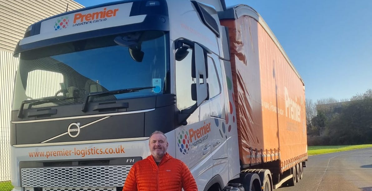 A man in an orange jacket standing in front of an orange Premier Logistics truck