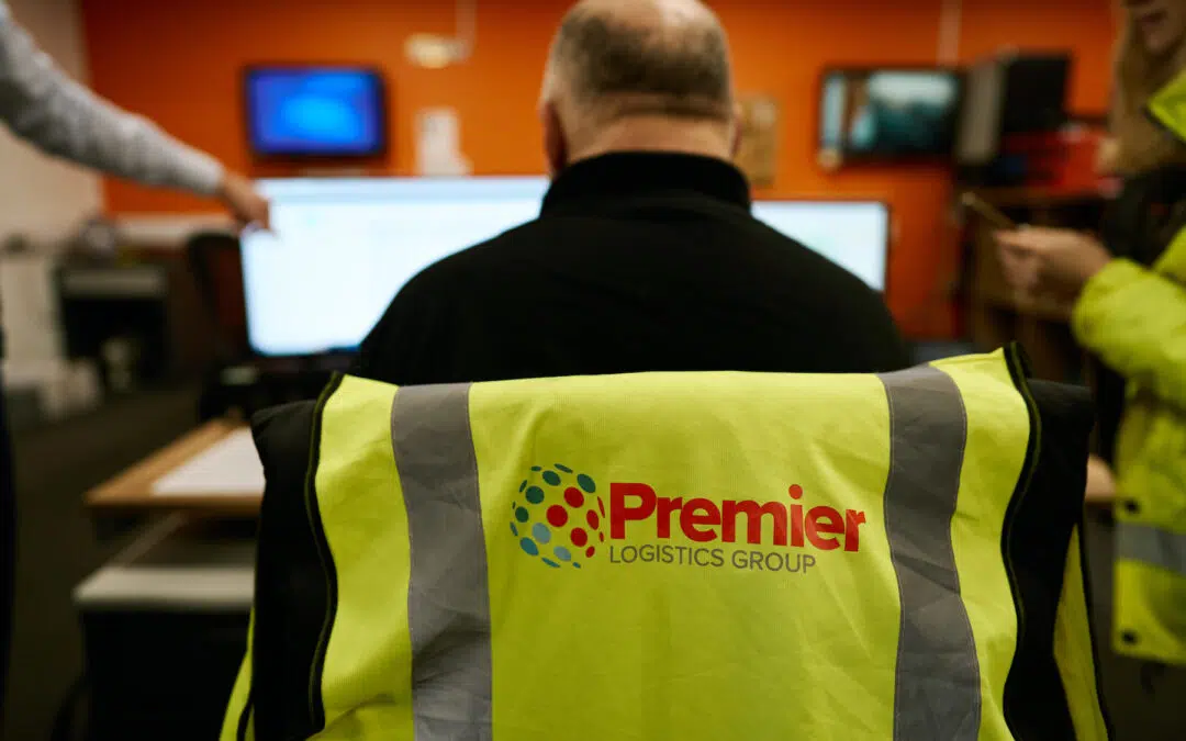 a Premier Logistics hi vis vest over a chair while a man works on a computer