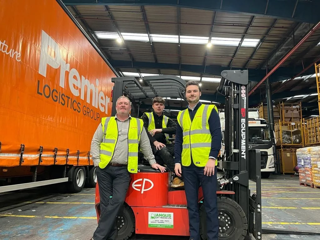 Three men with high vis jackets sitting around a forklift in a warehouse next to an orange lorry